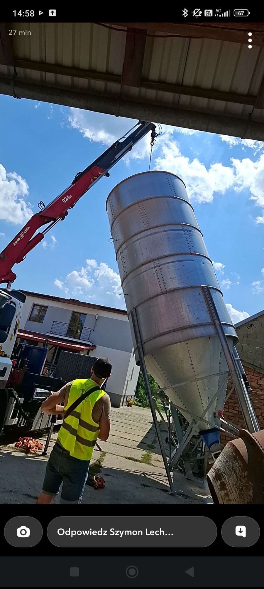 Dźwig transportuje metalowy silos w Tarnawie. Widoczny pracownik w kamizelce odblaskowej i fragment budynku w tle. Słoneczny dzień, niebieskie niebo.
