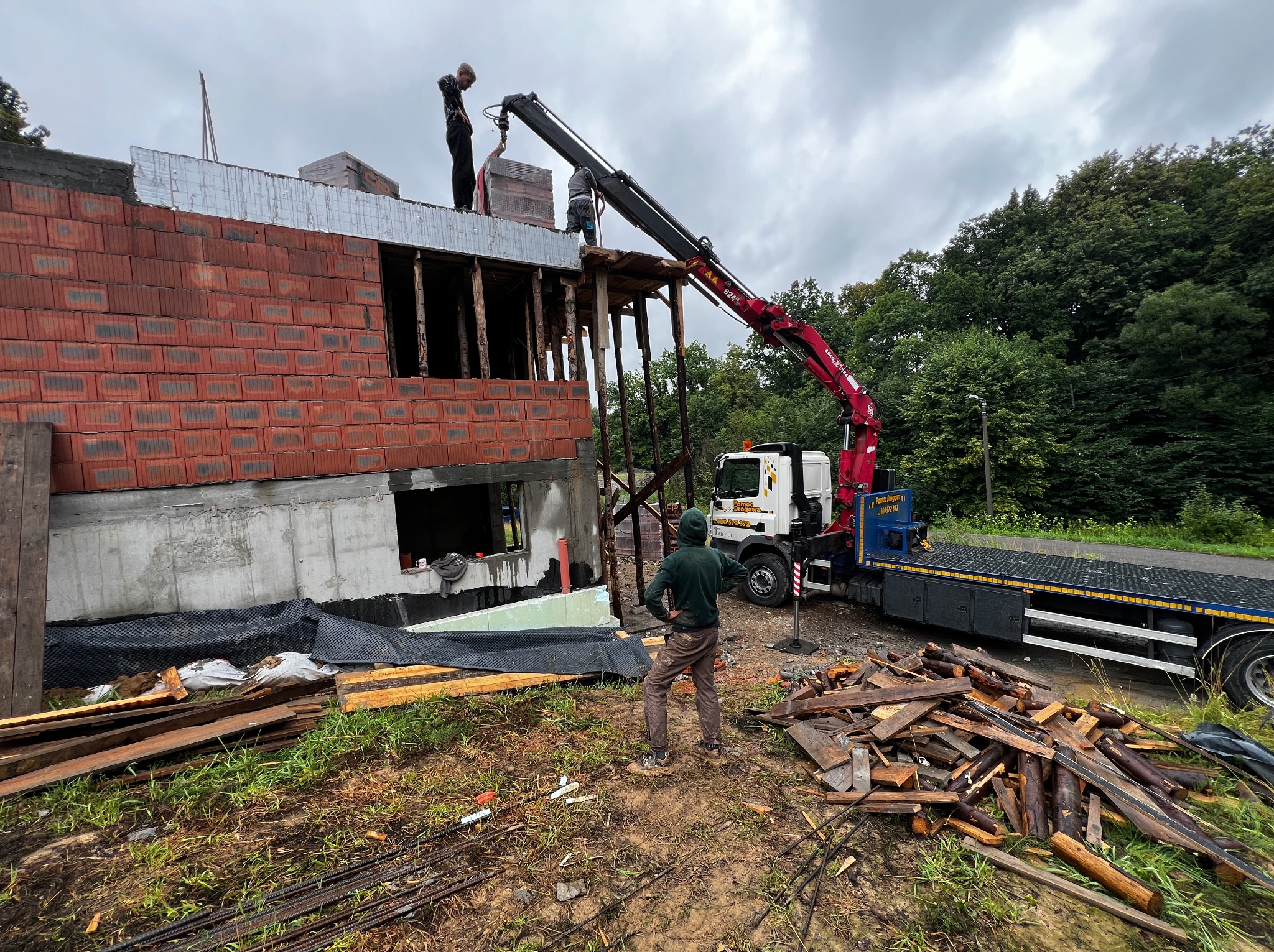 Budowa domu z czerwonej cegły. Żuraw przenosi materiały na górę. Widoczny pracownik w zielonej bluzie i laweta. Tło: las i pochmurne niebo.