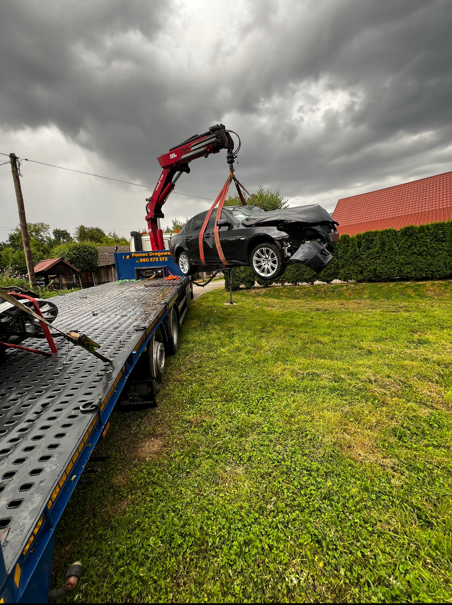 Czarny, uszkodzony samochód podnoszony przez czerwony dźwig lawety na tle pochmurnego nieba i zielonego trawnika. Widoczna część platformy lawety.