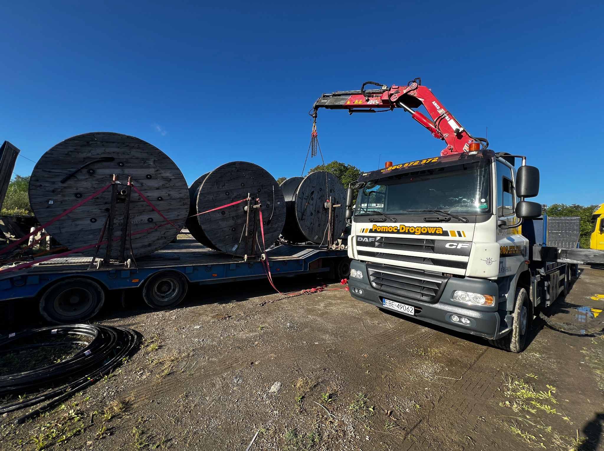 Laweta z dźwigiem transportuje duże, drewniane szpule z kablami. Widoczny numer telefonu na kabinie. Błękitne niebo w tle. Transport specjalistyczny.