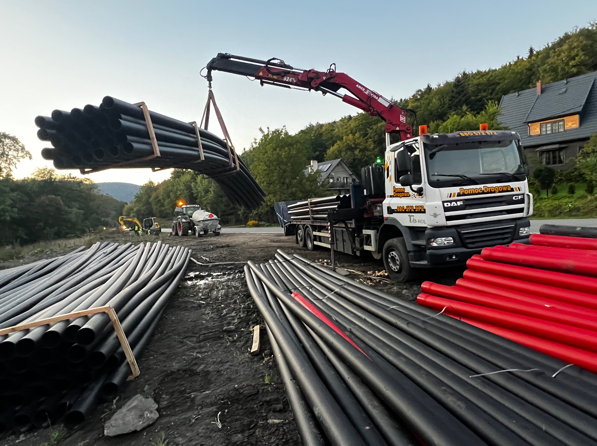 Ciężarówka z dźwigiem transportuje rury na tle zieleni i budynku. Na pierwszym planie stosy czarnych i czerwonych rur. Widoczny sprzęt budowlany w oddali.