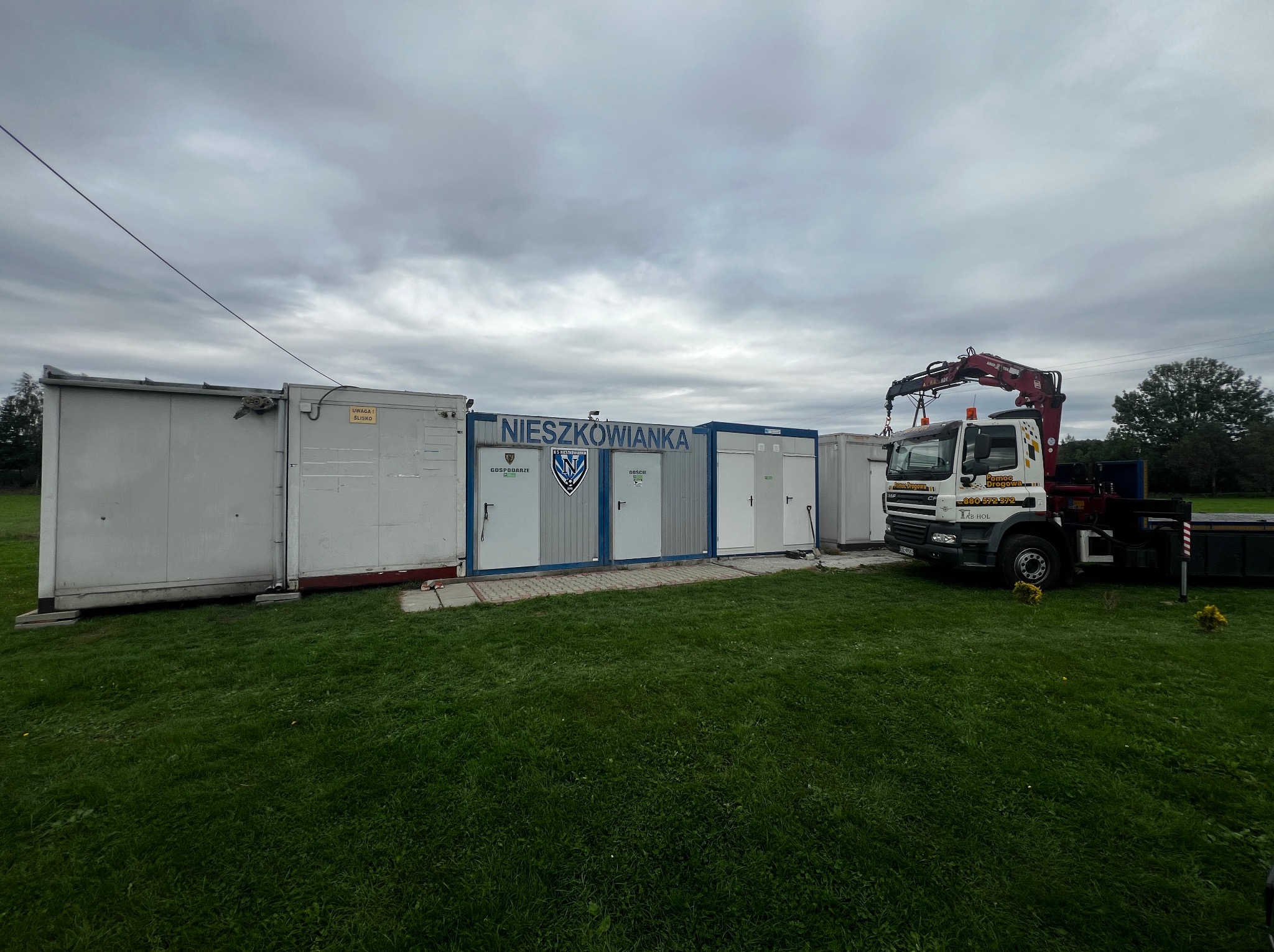 Ustawione kontenery z napisem 'Nieszkowianka' i zaparkowany obok nich samochód ciężarowy z dźwigiem na trawiastym terenie pod pochmurnym niebem.