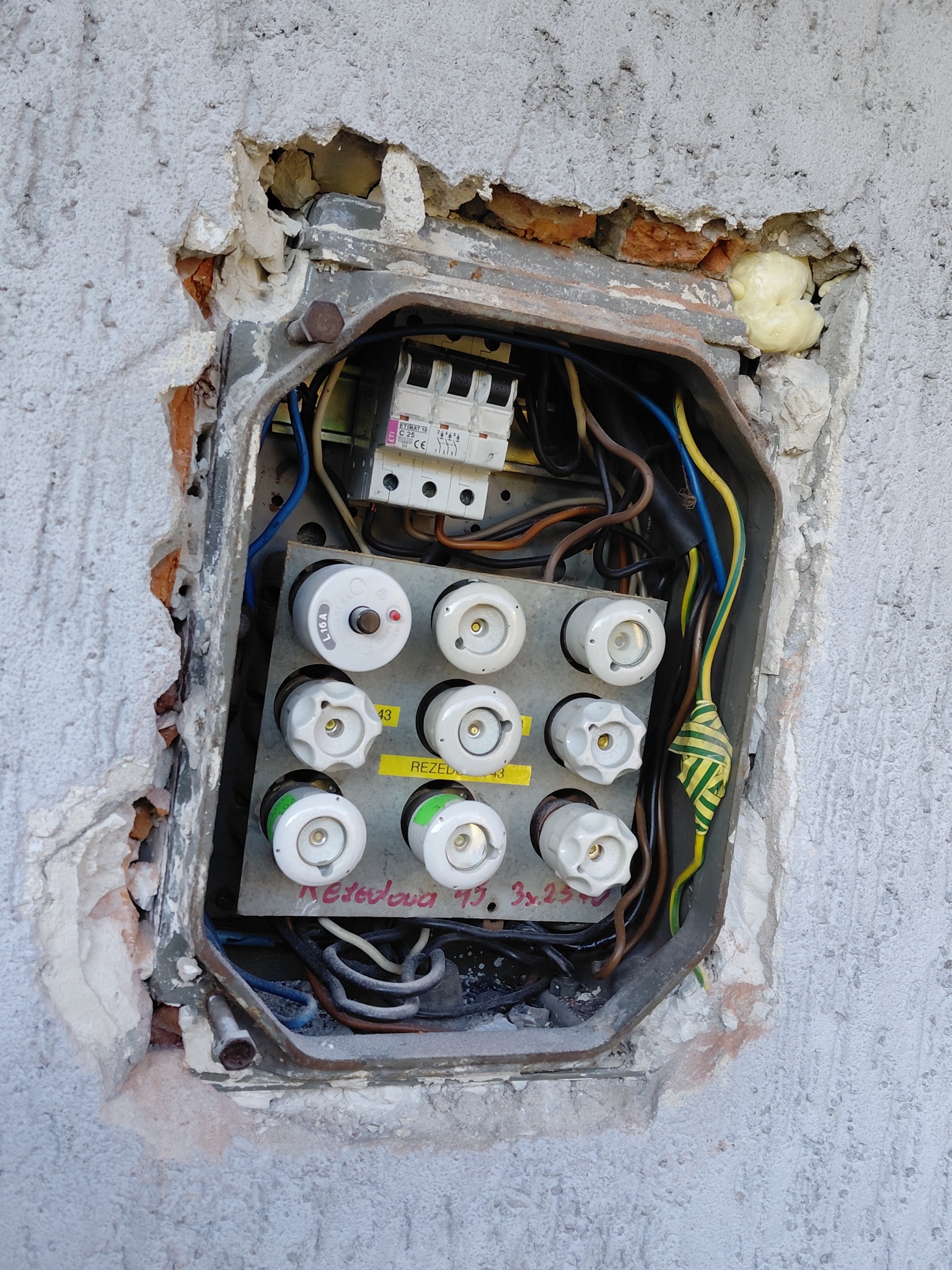 Otwarta skrzynka bezpiecznikowa w ścianie z widocznymi bezpiecznikami, przewodami i elementami instalacji. Widoczne uszkodzenia ściany wokół skrzynki.