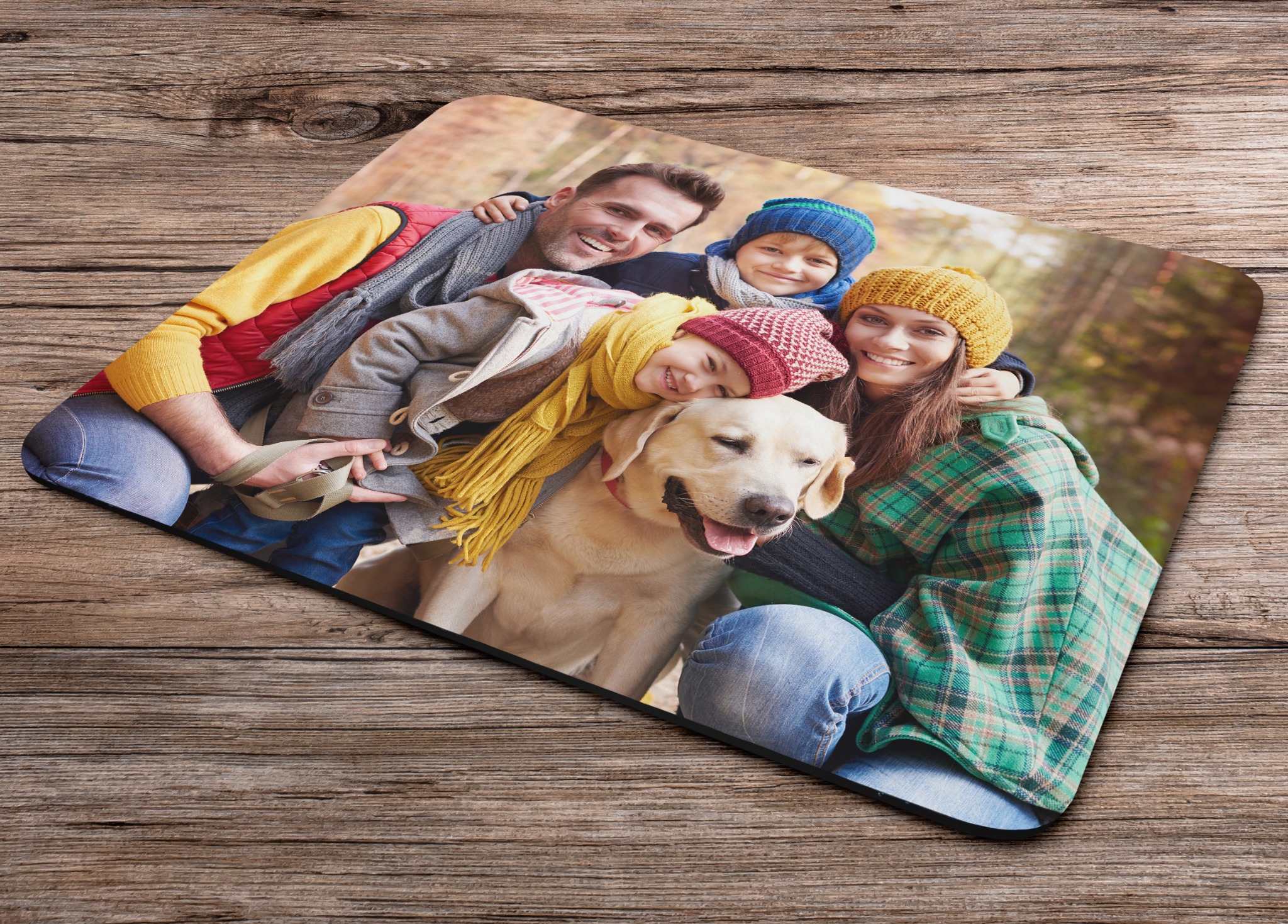 Podkładka pod mysz z nadrukiem: uśmiechnięta rodzina z psem na tle jesiennego lasu. Żywe kolory, wyraźny nadruk, położona na drewnianym stole.