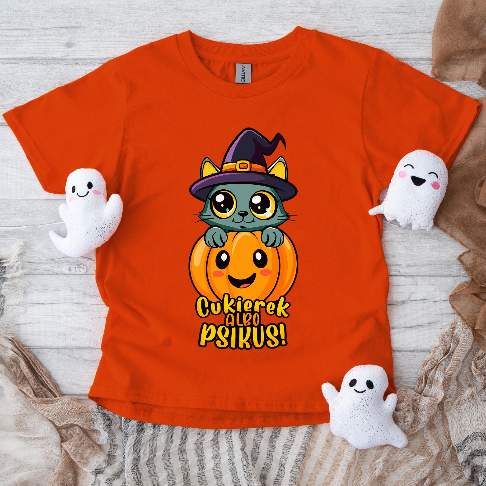 Pomarańczowa koszulka z halloweenowym nadrukiem kota w kapeluszu na dyni, otoczona pluszowymi duszkami, leży na drewnianym tle z tkaniną.