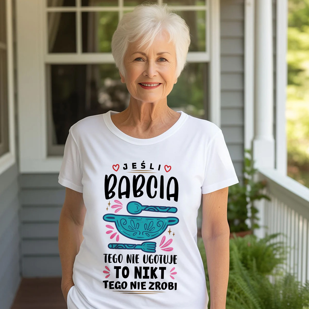 Uśmiechnięta seniorka w białej koszulce z nadrukiem 'Jeśli Babcia tego nie ugotuje to nikt tego nie zrobi' na tle werandy z kolumnami.