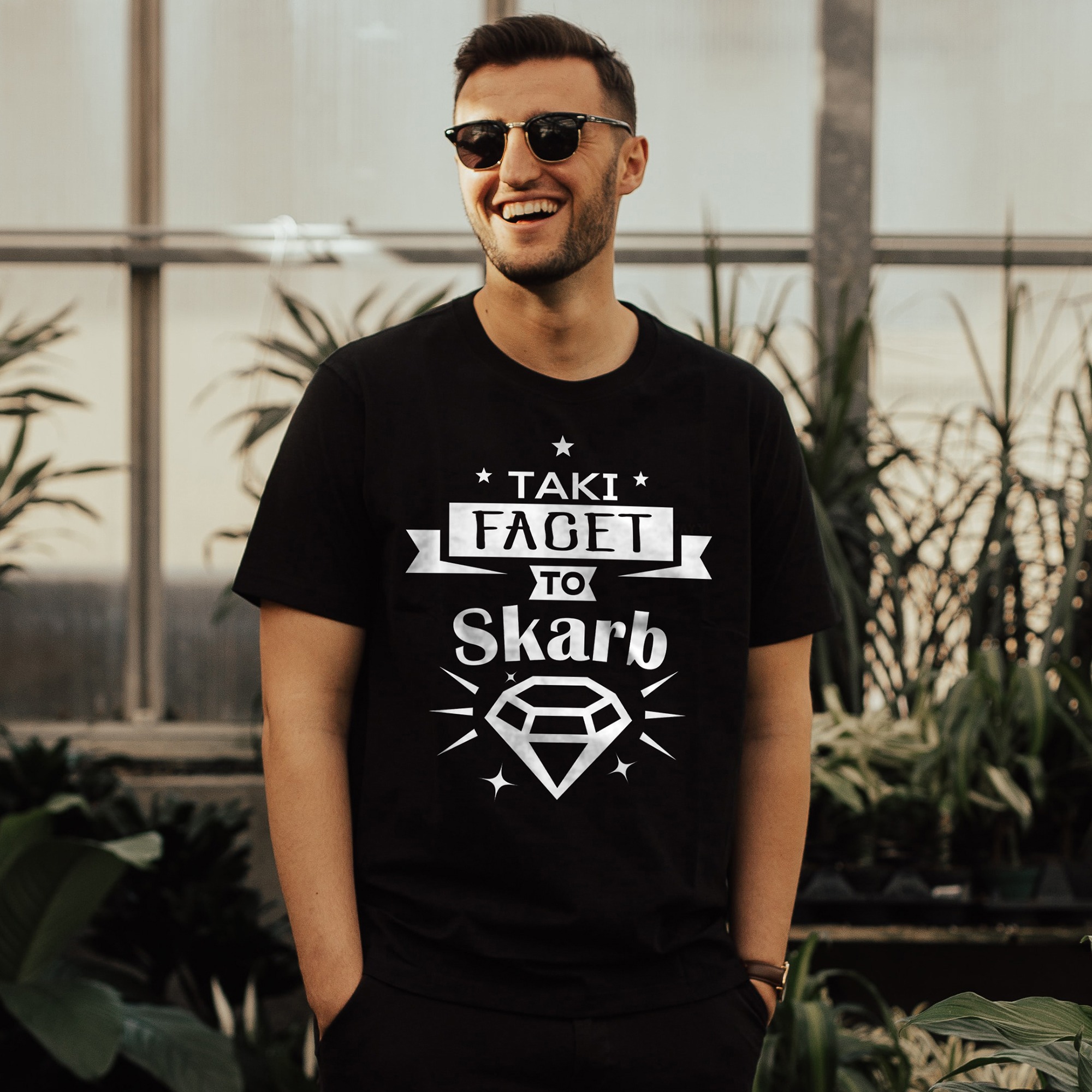 Uśmiechnięty mężczyzna w okularach, ubrany w czarny T-shirt z białym nadrukiem 'Taki facet to Skarb' i motywem diamentu, na tle szklarni z roślinami.