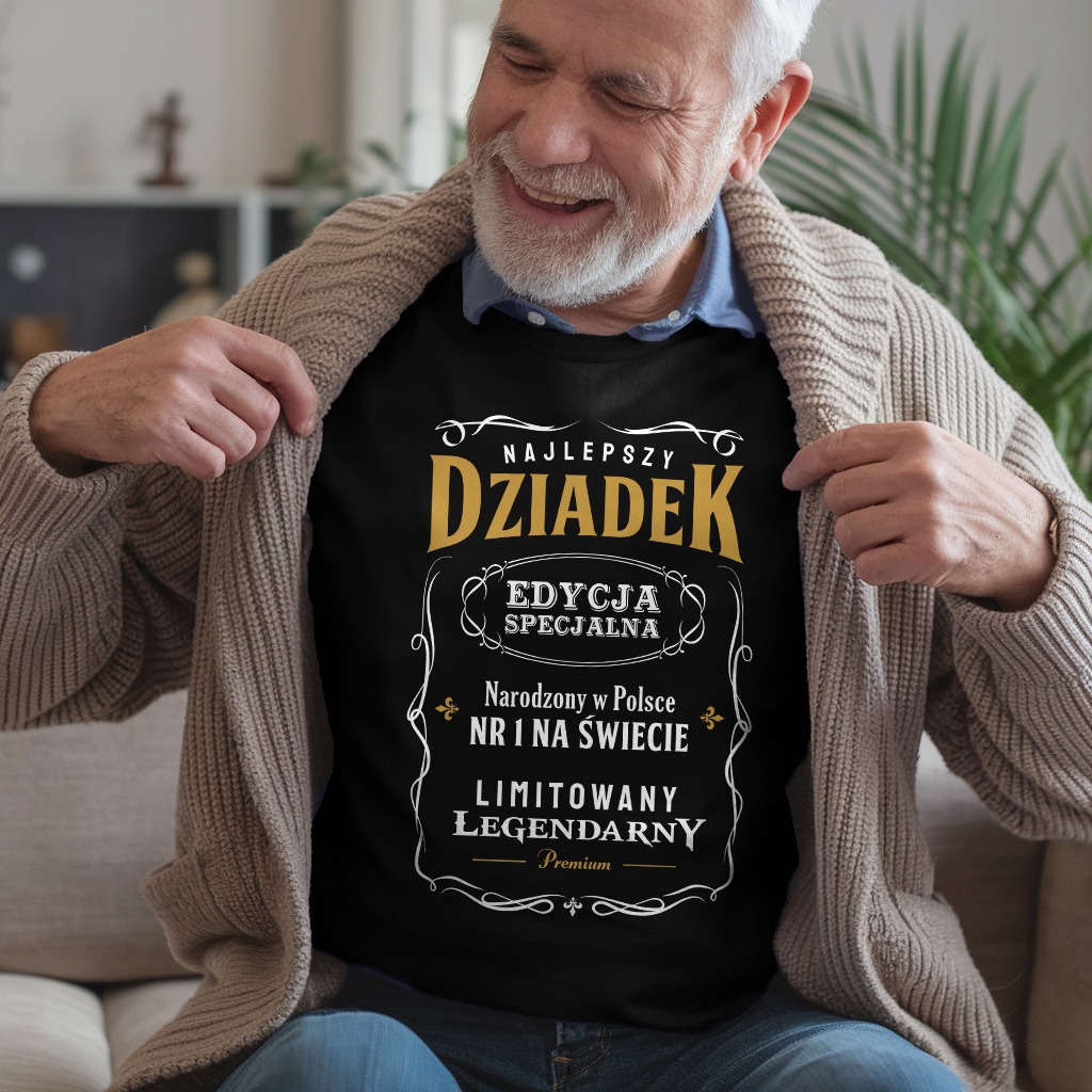 Uśmiechnięty starszy mężczyzna prezentuje czarną koszulkę z napisem 'Najlepszy Dziadek' w ozdobnej grafice, siedząc na kanapie w jasnym swetrze.