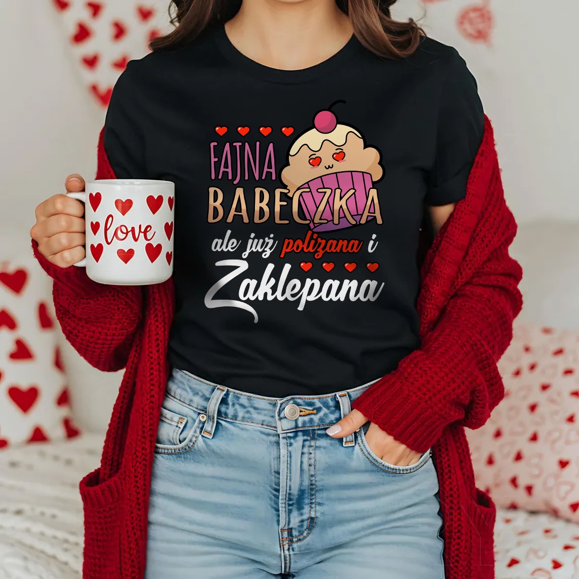 Kobieta w czarnej koszulce z nadrukiem 'Fajna Babeczka' i graficznym babeczką trzyma kubek z sercami i napisem 'love', ubrana w czerwony kardigan.