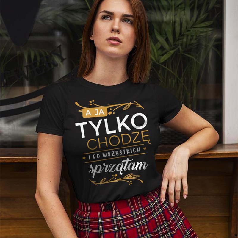 Kobieta prezentuje czarną koszulkę z napisem „A ja tylko chodzę i po wszystkich sprzątam” na tle roślin i drewnianego blatu. Ujęcie z delikatnym oświetleniem.