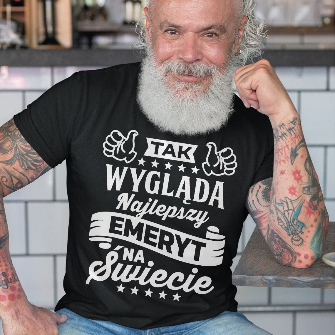 Mężczyzna z brodą w czarnej koszulce z białym nadrukiem 'Tak wygląda najlepszy emeryt na świecie', siedzący przy stole. Widoczne tatuaże na rękach.