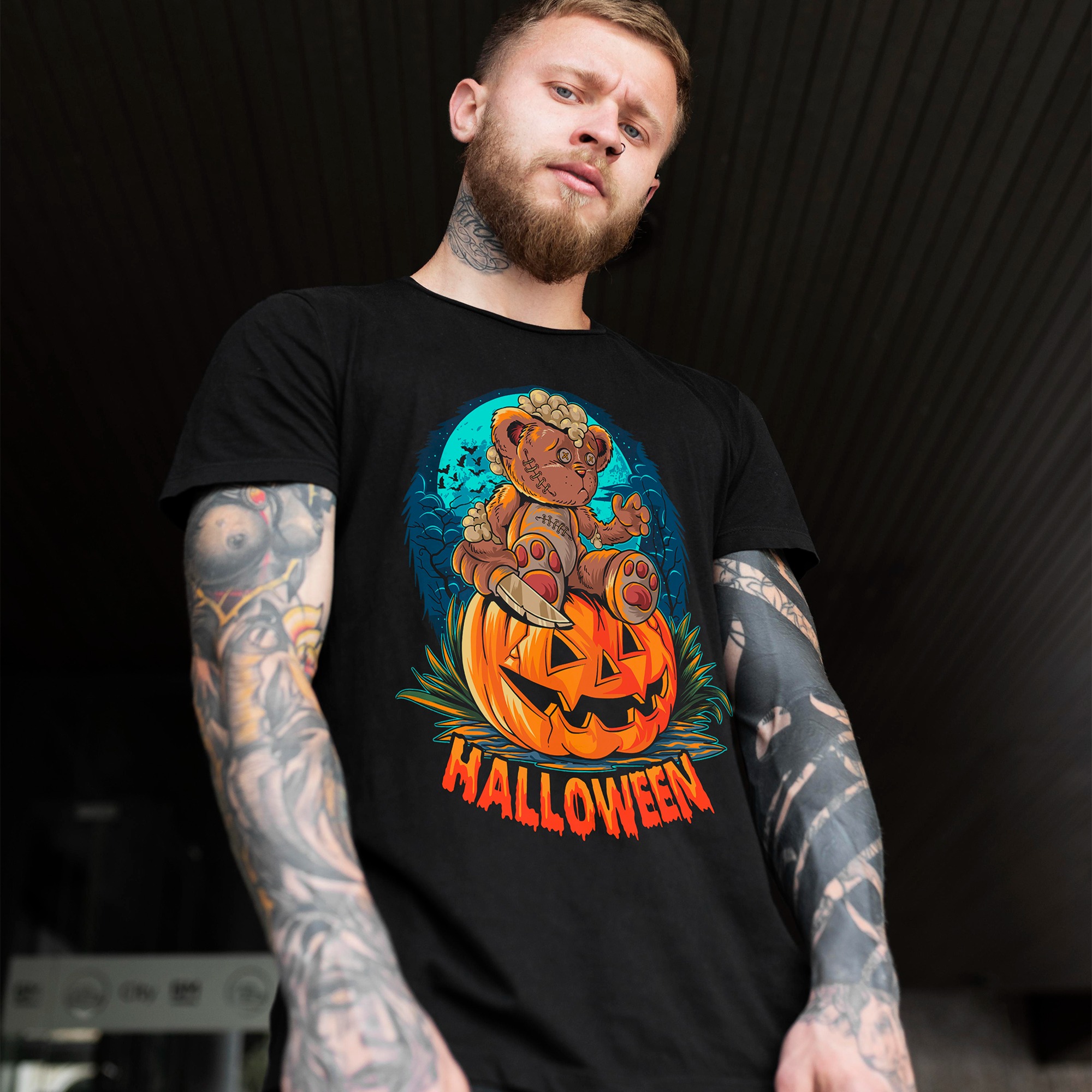 Mężczyzna z tatuażami prezentuje czarną koszulkę z nadrukiem misia zombie na dyni z napisem Halloween. Ujęcie z dołu, ciemne tło.