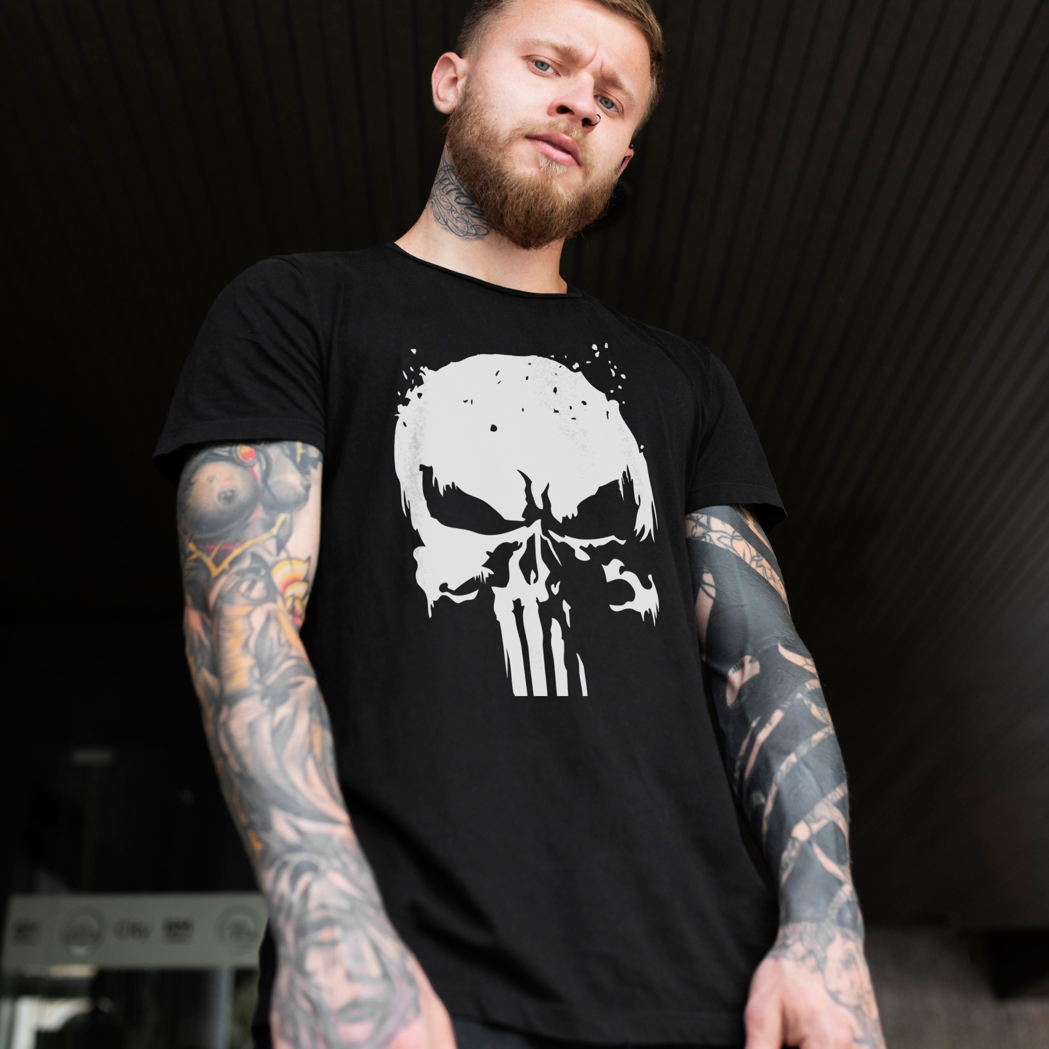 Mężczyzna z brodą i tatuażami prezentuje czarną koszulkę z białym nadrukiem czaszki na tle ciemnej ściany. Ujęcie z dołu.