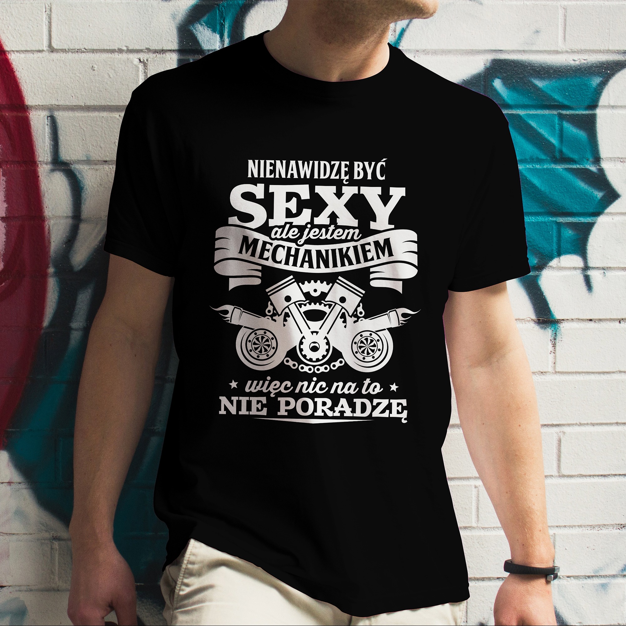 Mężczyzna w czarnym T-shircie z białym nadrukiem: 'Nienawidzę być SEXY ale jestem MECHANIKIEM, więc nic na to nie poradzę'. Tło to mur z graffiti.