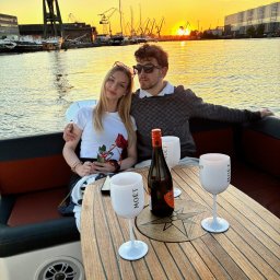 MS-LOGISTYKA MARIUSZ SOBAŃSKI - Para na jachcie przy zachodzie słońca, butelka i kieliszki Moët na drewnianym stole. W tle dźwigi portowe i budynki. Romantyczna atmosfera.