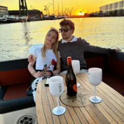 Para na jachcie przy zachodzie słońca, butelka i kieliszki Moët na drewnianym stole. W tle dźwigi portowe i budynki. Romantyczna atmosfera.