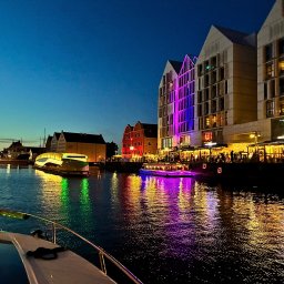 MS-LOGISTYKA MARIUSZ SOBAŃSKI - Nocny widok na gdańskie nabrzeże z pokładu łodzi, odbijające się w wodzie kolorowe światła budynków i statków. Oświetlona restauracja wzdłuż brzegu.