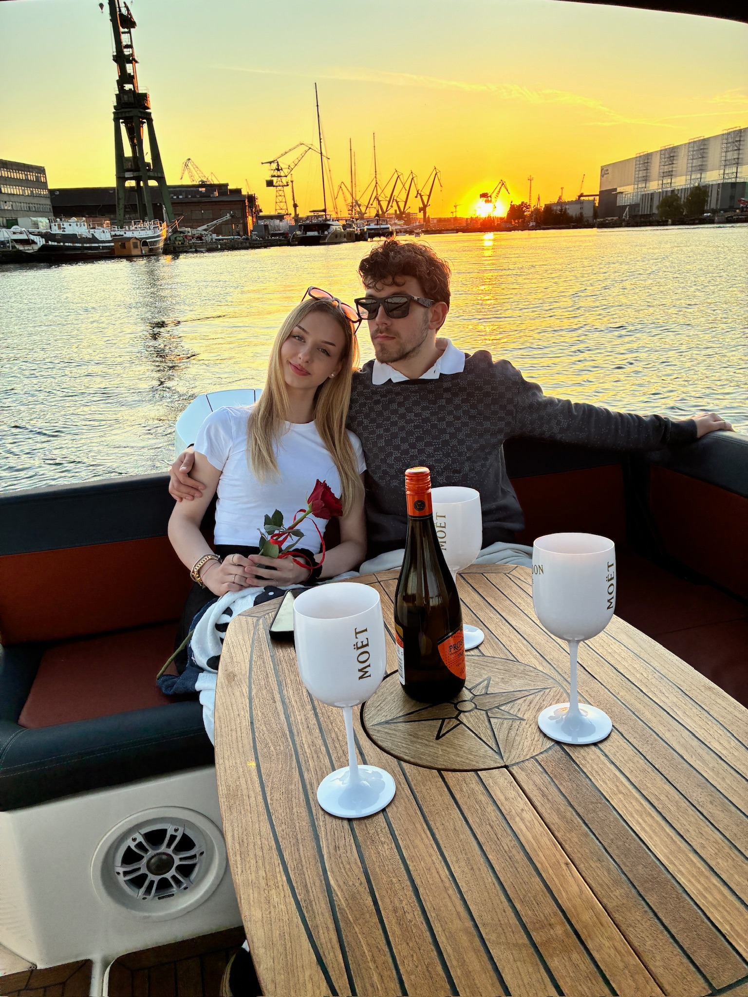 Para na jachcie przy zachodzie słońca, butelka i kieliszki Moët na drewnianym stole. W tle dźwigi portowe i budynki. Romantyczna atmosfera.