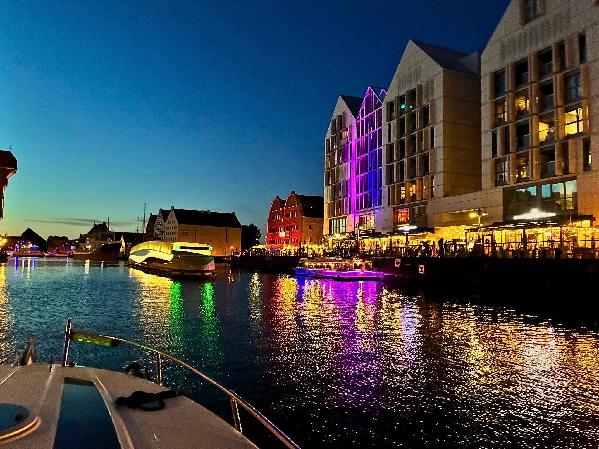 Nocny widok na gdańskie nabrzeże z pokładu łodzi, odbijające się w wodzie kolorowe światła budynków i statków. Oświetlona restauracja wzdłuż brzegu.