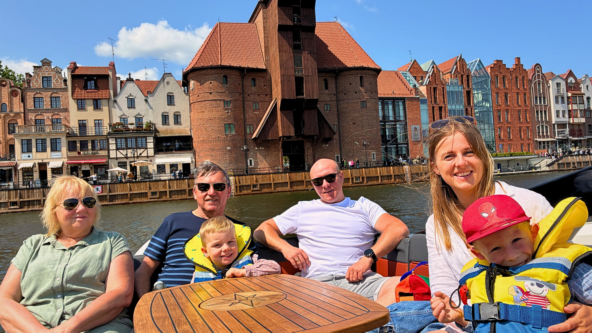 Rodzinny rejs po Motławie w Gdańsku. Grupa osób na łodzi z Żurawiem Gdańskim w tle. Słoneczny dzień, błękitne niebo. Wszyscy uśmiechnięci, relaks na wodzie.