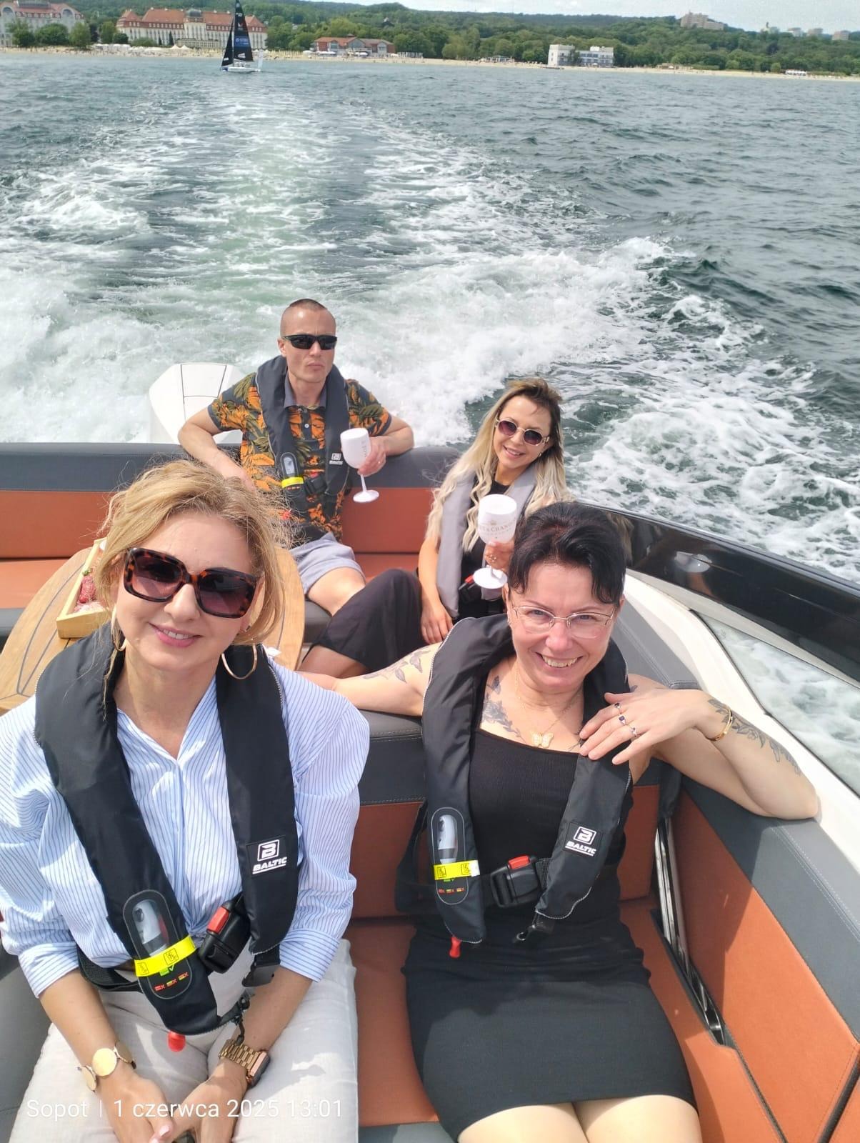 Grupa osób w kamizelkach na motorówce, w tle morze i budynki. Uczestnicy wycieczki z kieliszkami w dłoniach, relaksują się podczas rejsu.