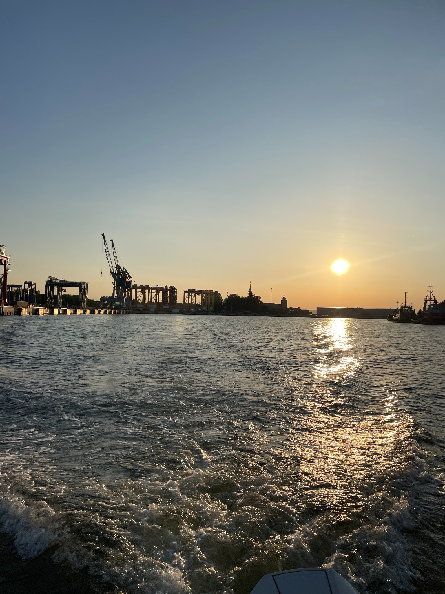 Widok na port z perspektywy łodzi motorowej podczas zachodu słońca. Woda wzburzona, ślady po śrubie. Na horyzoncie dźwigi portowe.