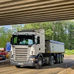 Ciężarówka Scania z naczepą pod betonowym wiaduktem, w tle zieleń drzew. Na drodze rozłożony materiał sypki. Widoczne detale pojazdu i otoczenia.