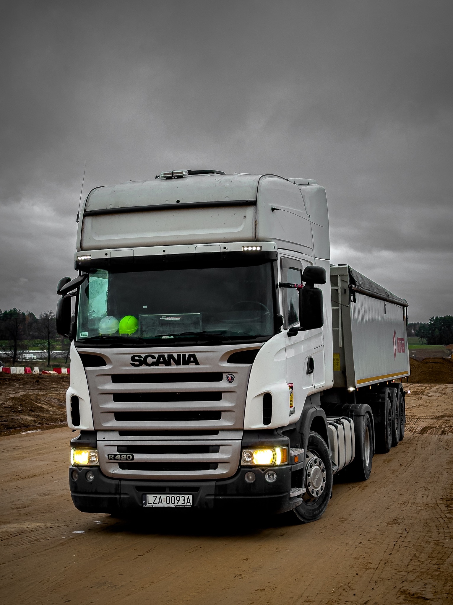 Ciężarówka Scania R420 na placu budowy, przód pojazdu z włączonymi światłami, widoczne kaski ochronne na desce rozdzielczej, pochmurne niebo w tle.