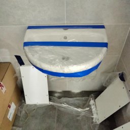 AD BUD USŁUGI Remontowo Budowlane - Biała umywalka zabezpieczona folią i niebieską taśmą malarską, przygotowana do montażu. Obok widoczne elementy szafki pod umywalkę i kartonowe pudło.