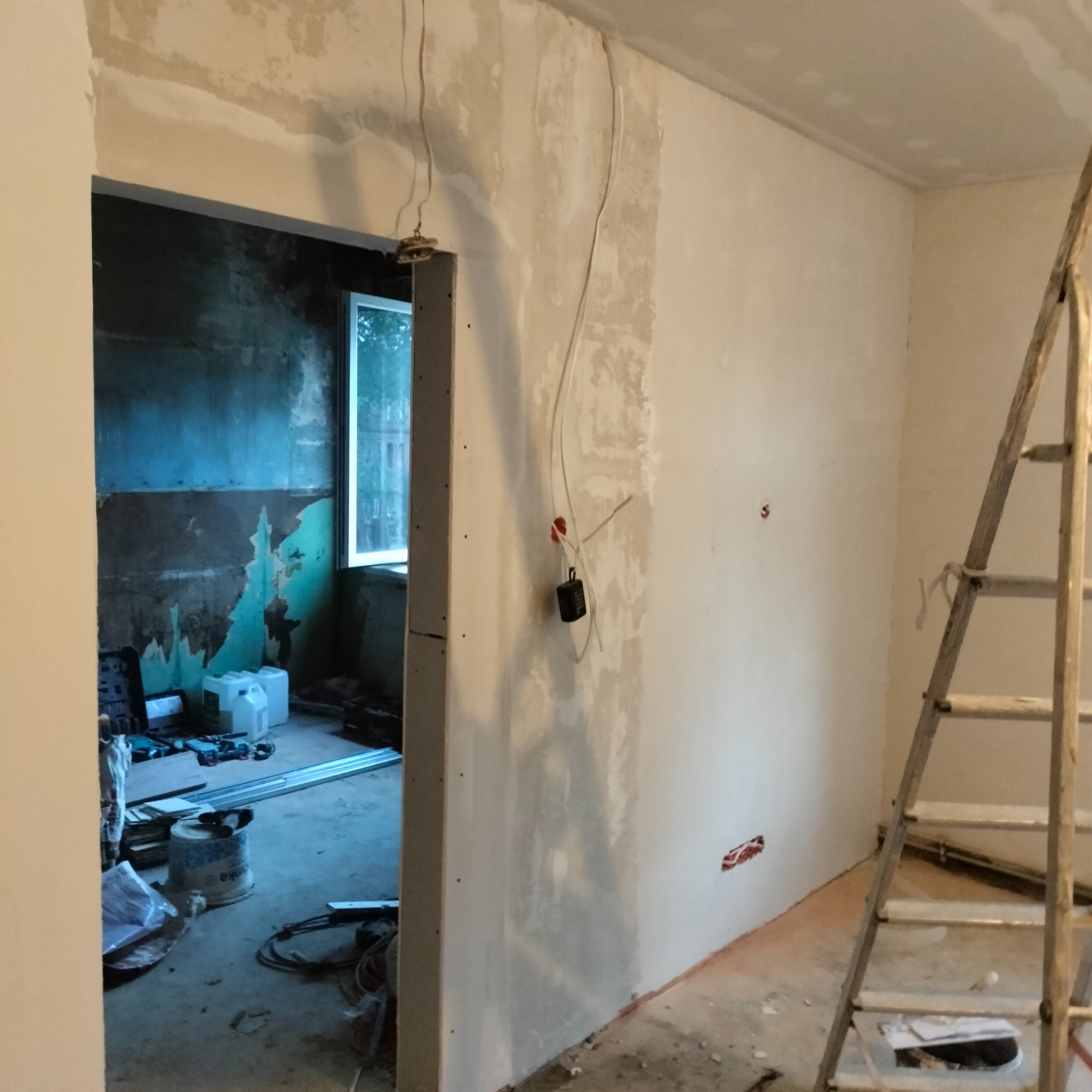 Wnętrze w trakcie remontu: fragment ściany z widocznymi przewodami elektrycznymi, fragmentem futryny drzwiowej i drabiną. W tle widoczne narzędzia i materiały budowlane.