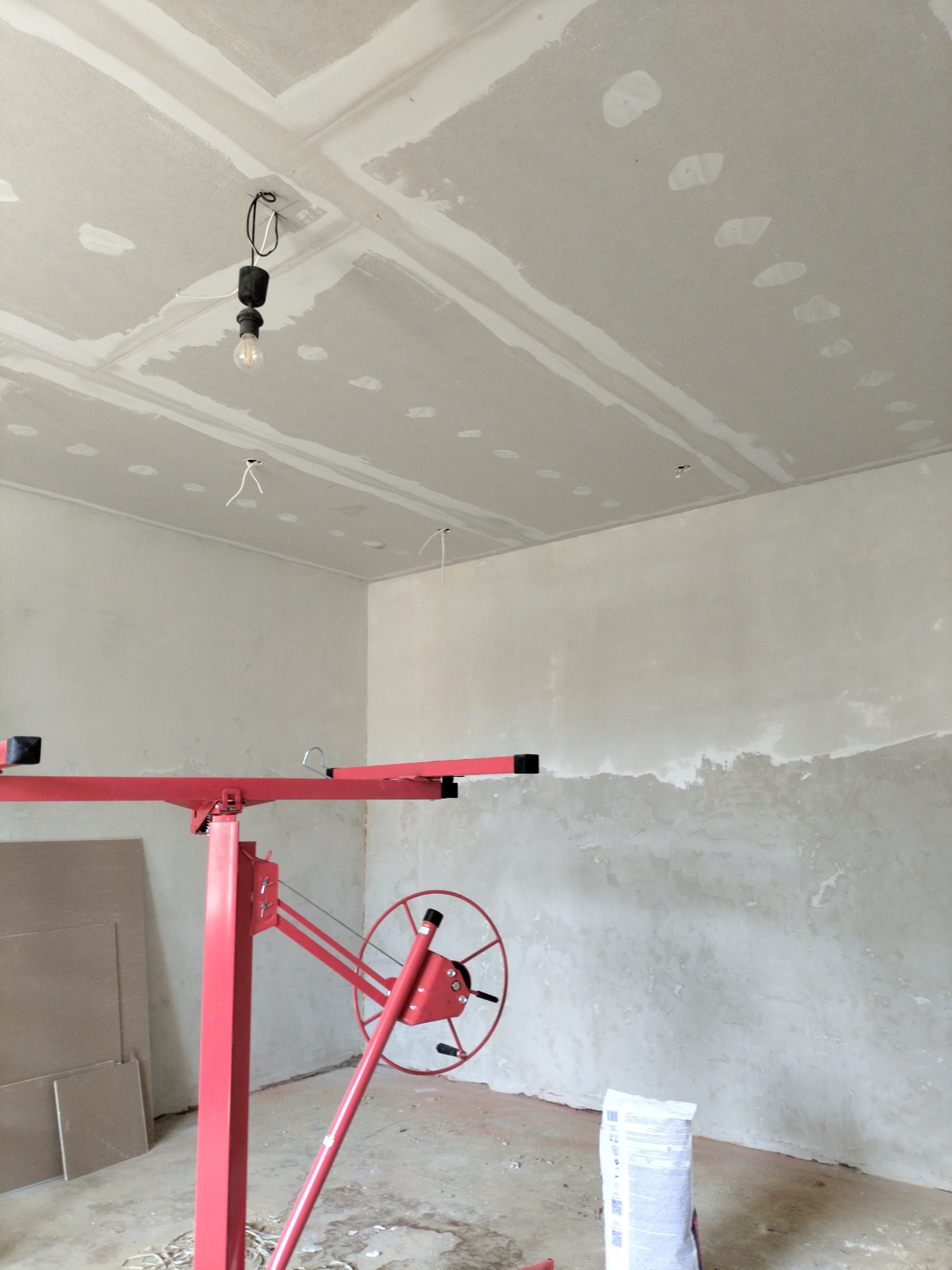 Wnętrze w trakcie wykończenia: sufit z płyt gipsowo-kartonowych z widocznymi łączeniami, czerwony podnośnik do płyt i worek gładzi szpachlowej.