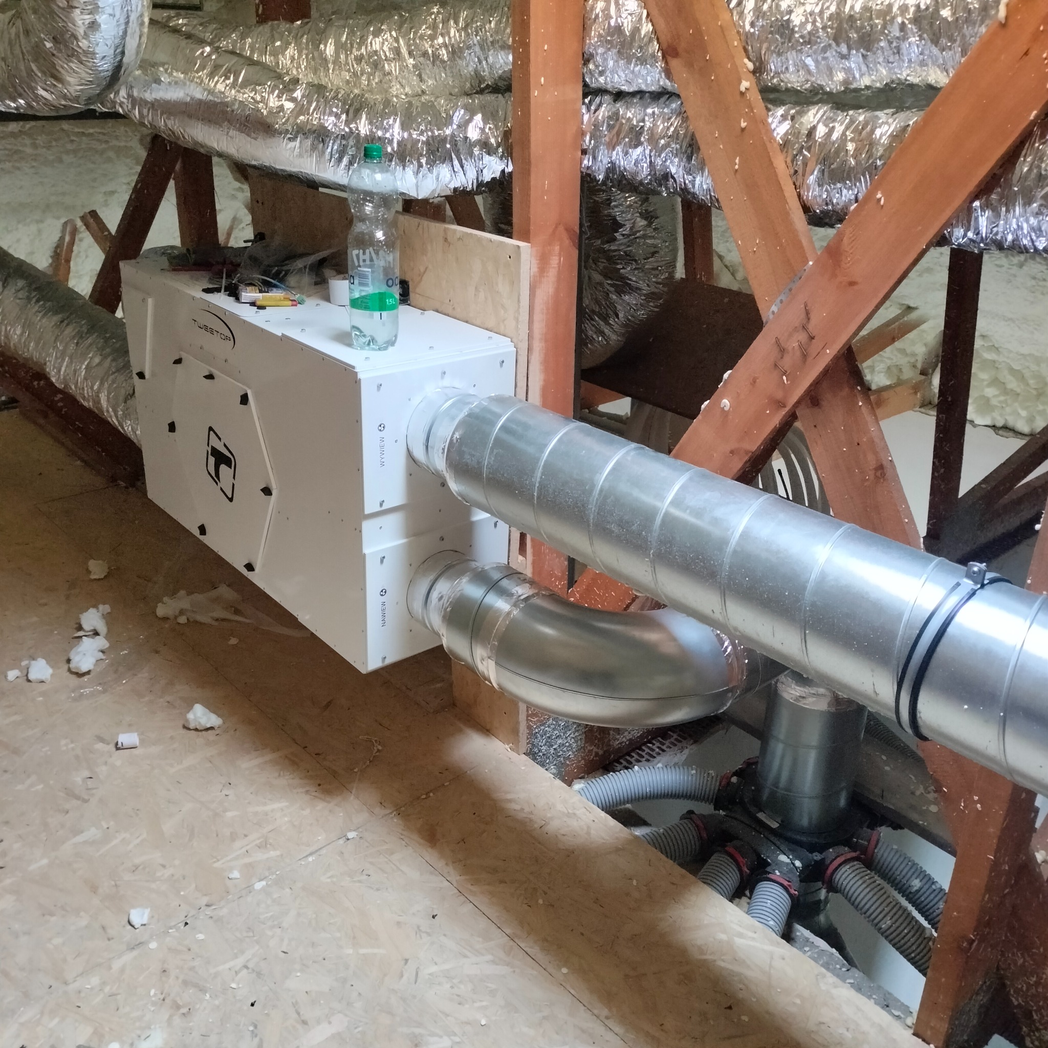 Biała jednostka wentylacyjna z podłączonymi metalowymi rurami w przestrzeni poddasza, widoczne drewniane elementy konstrukcyjne i izolacja.