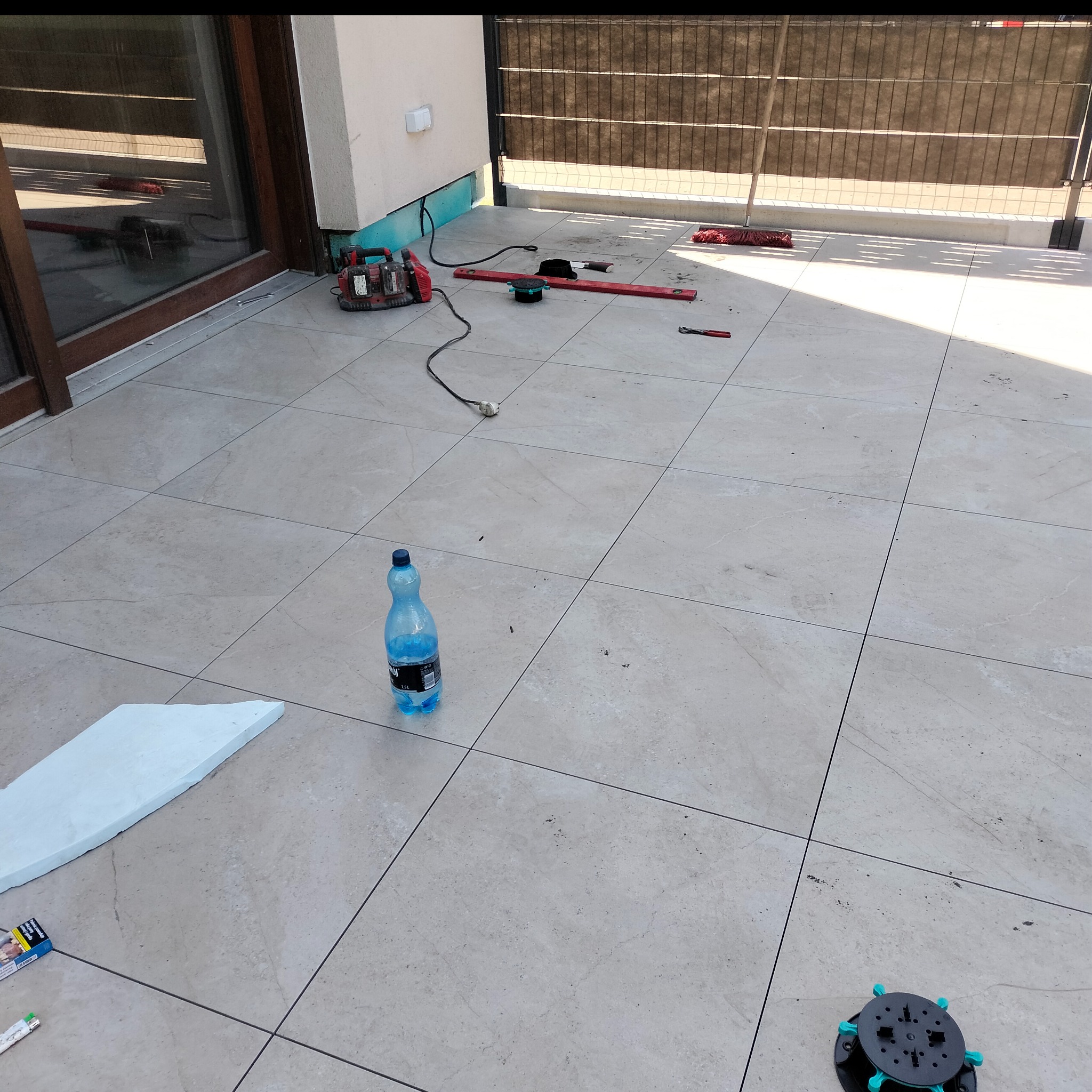 Układanie płytek na balkonie: poziomica, narzędzia, maszyna do cięcia, butelka wody. Widoczny etap prac wykończeniowych na zewnątrz budynku.