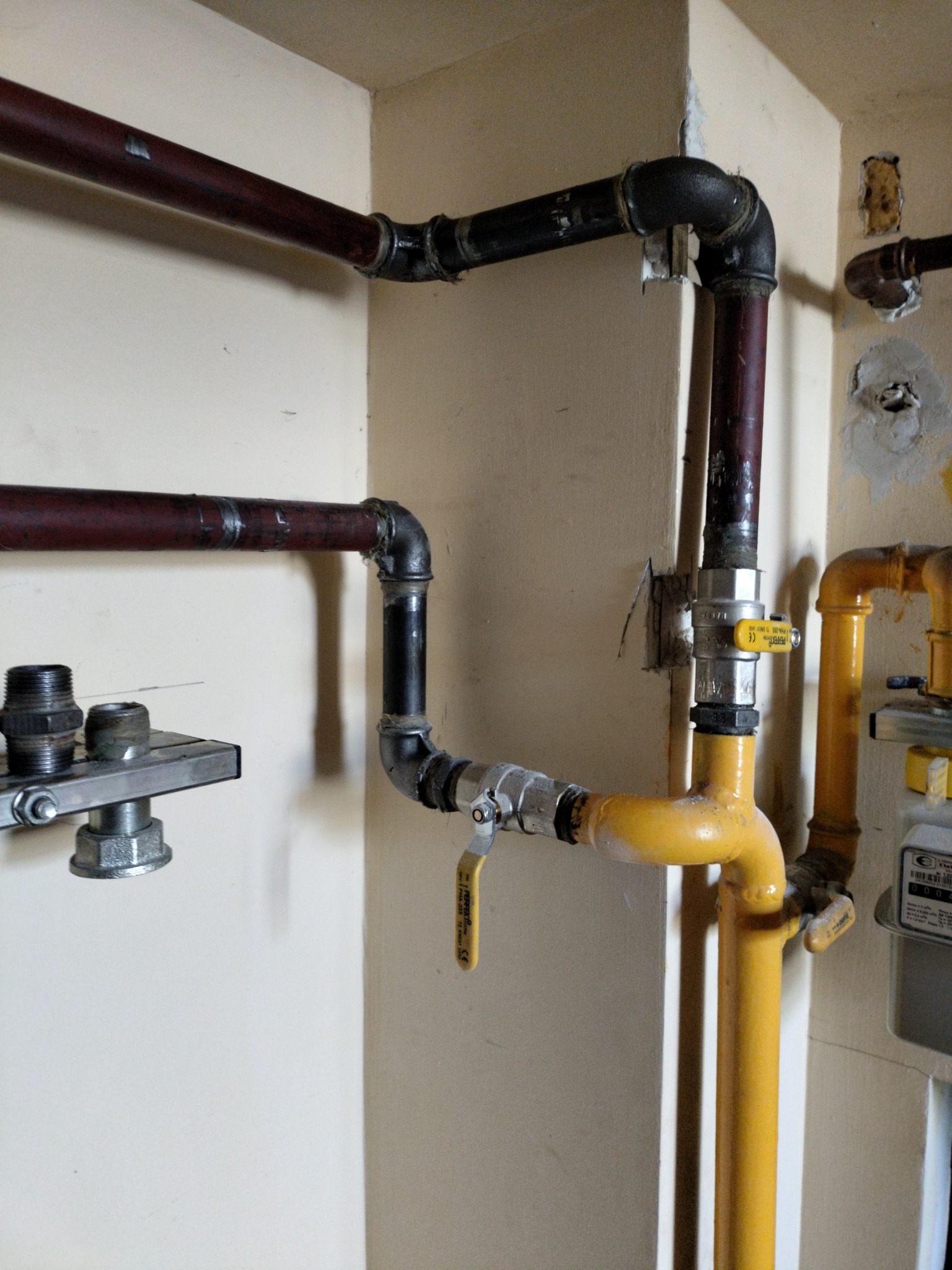 Wnętrze z fragmentem instalacji gazowej: żółte i ciemne rury, zawór z żółtą rączką, złączki i elementy montażowe przy ścianie. Widoczne ślady po ingerencji w ścianę.