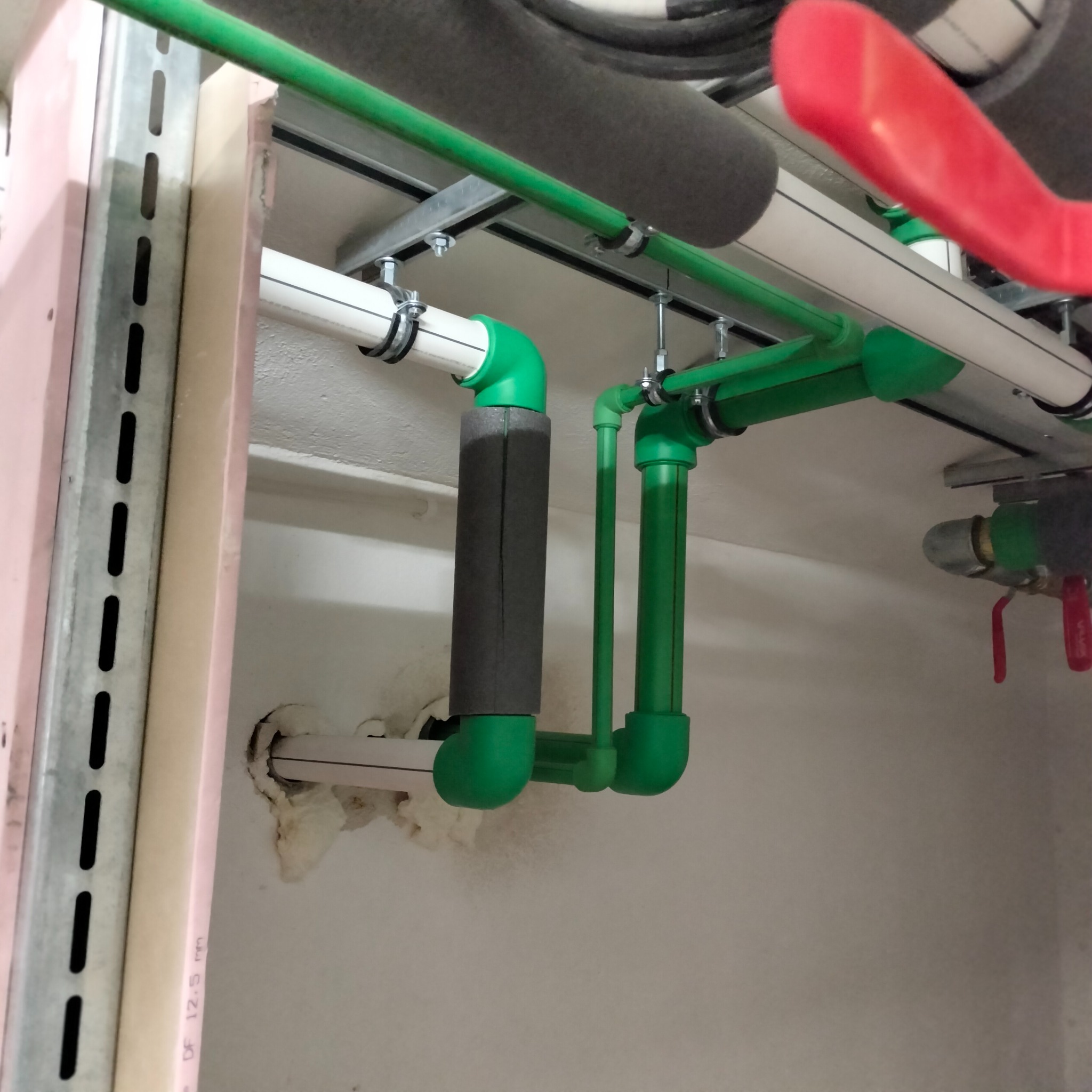 Fragment instalacji hydraulicznej z zielonymi rurami PPr, zaworami i izolacją termiczną, zamocowanej pod sufitem. Widoczne elementy konstrukcyjne i ściana.