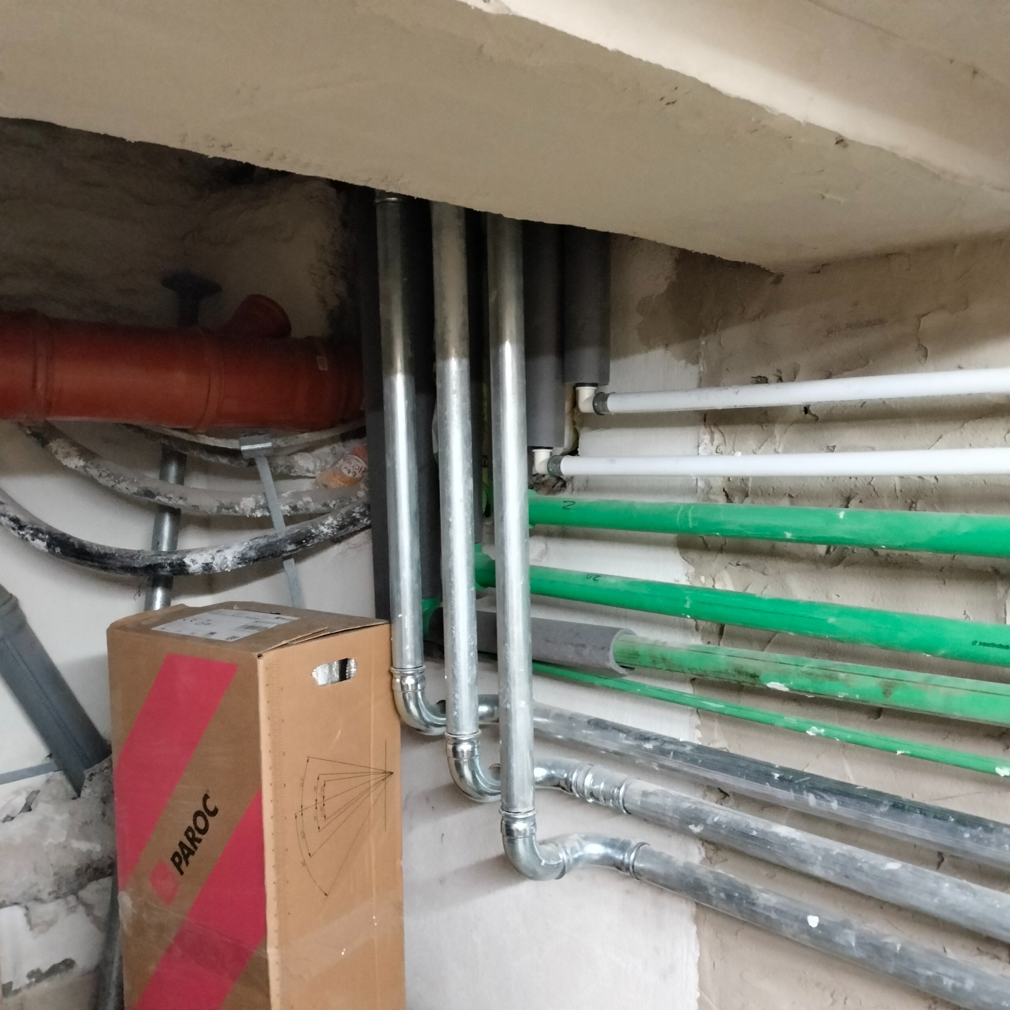 Różnokolorowe rury instalacyjne (stalowe, plastikowe) ułożone wzdłuż ściany pod sufitem. Widoczny karton z napisem PAROC.
