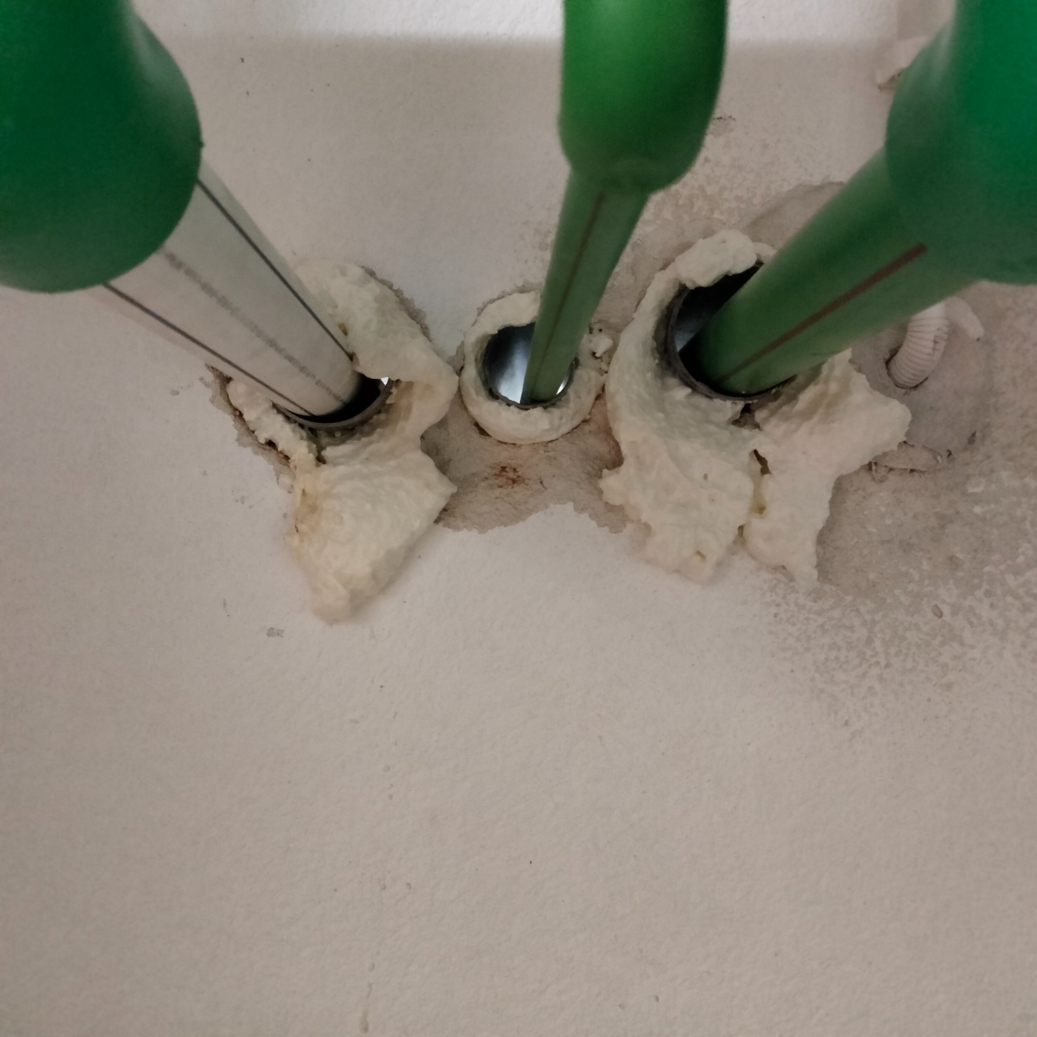 Świeżo uszczelnione pianką montażową, zielone rury instalacyjne wystające ze ściany. Widoczne otwory w ścianie i fragment białej instalacji elektrycznej.