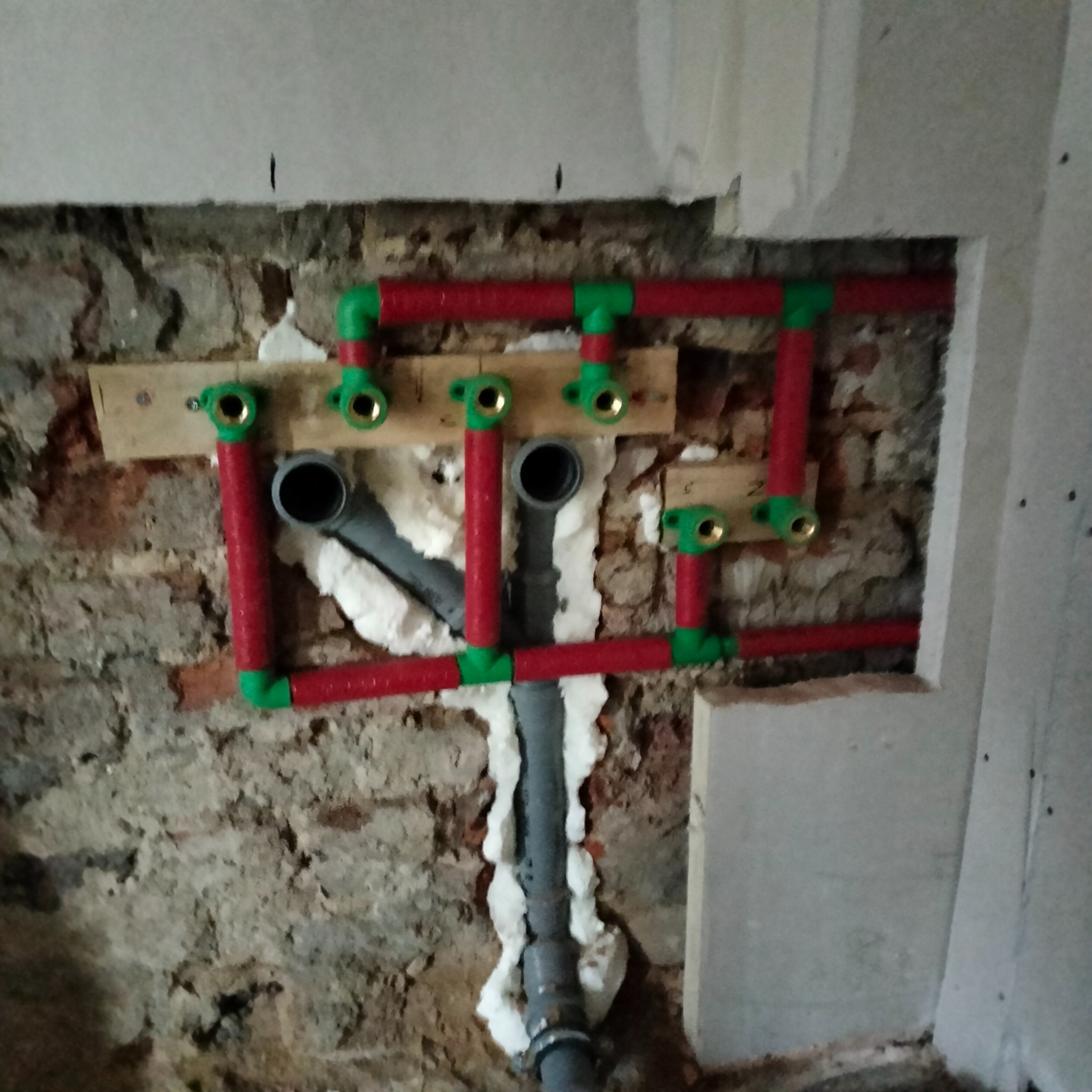 Czerwono-zielona instalacja wodna na tle surowej ściany z cegły i betonu, zabezpieczona pianką montażową. Widoczne mosiężne złączki i szare rury kanalizacyjne.