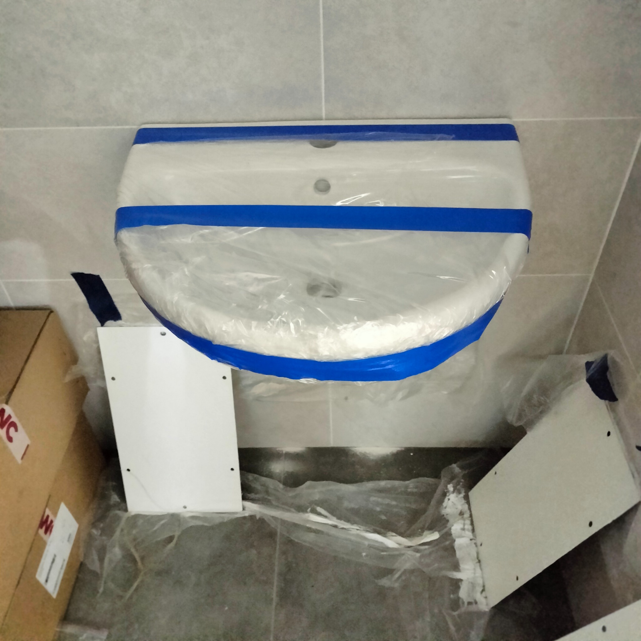 Biała umywalka zabezpieczona folią i niebieską taśmą malarską, przygotowana do montażu. Obok widoczne elementy szafki pod umywalkę i kartonowe pudło.