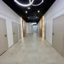 Długi korytarz biurowy z szarymi drzwiami i ceglanymi wstawkami, oświetlony nowoczesnymi lampami wiszącymi w kształcie okręgów. Ciemny sufit i jasna podłoga.