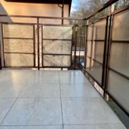 Jasne, ceramiczne płytki na balkonie z metalową balustradą i panelami z tworzywa. Widok na drzewa i budynek w tle. Słoneczny dzień.