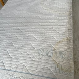 ORDEN - znaczy porządek - Materac z widocznymi plamami i zabrudzeniami na białym, pikowanym poszyciu. Widoczne napisy 'NATURA' i fragment 'SAFE'.