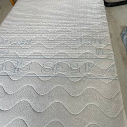 ORDEN - znaczy porządek - Biały materac z wzorem fal i napisem 'NATURAL CARE' oraz 'REGLIS SLEEP' w górnej części, leżący na podłodze, ukazujący strukturę i detale materiału.