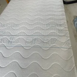 Biały materac z wzorem fal i napisem 'NATURAL CARE' oraz 'REGLIS SLEEP' w górnej części, leżący na podłodze, ukazujący strukturę i detale materiału.