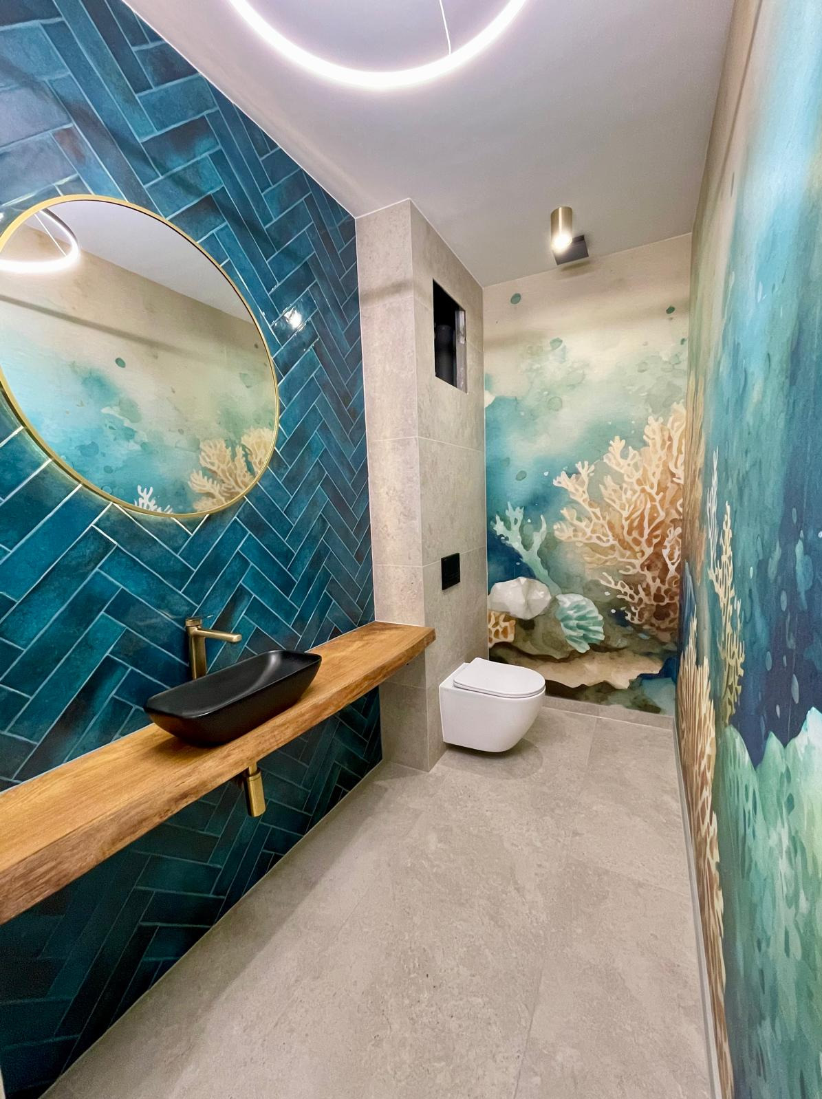 Wykończona łazienka po remoncie: turkusowa ściana z lustrem, drewniany blat z czarną umywalką, toaleta podwieszana, ściana z motywem morskim, szara podłoga.