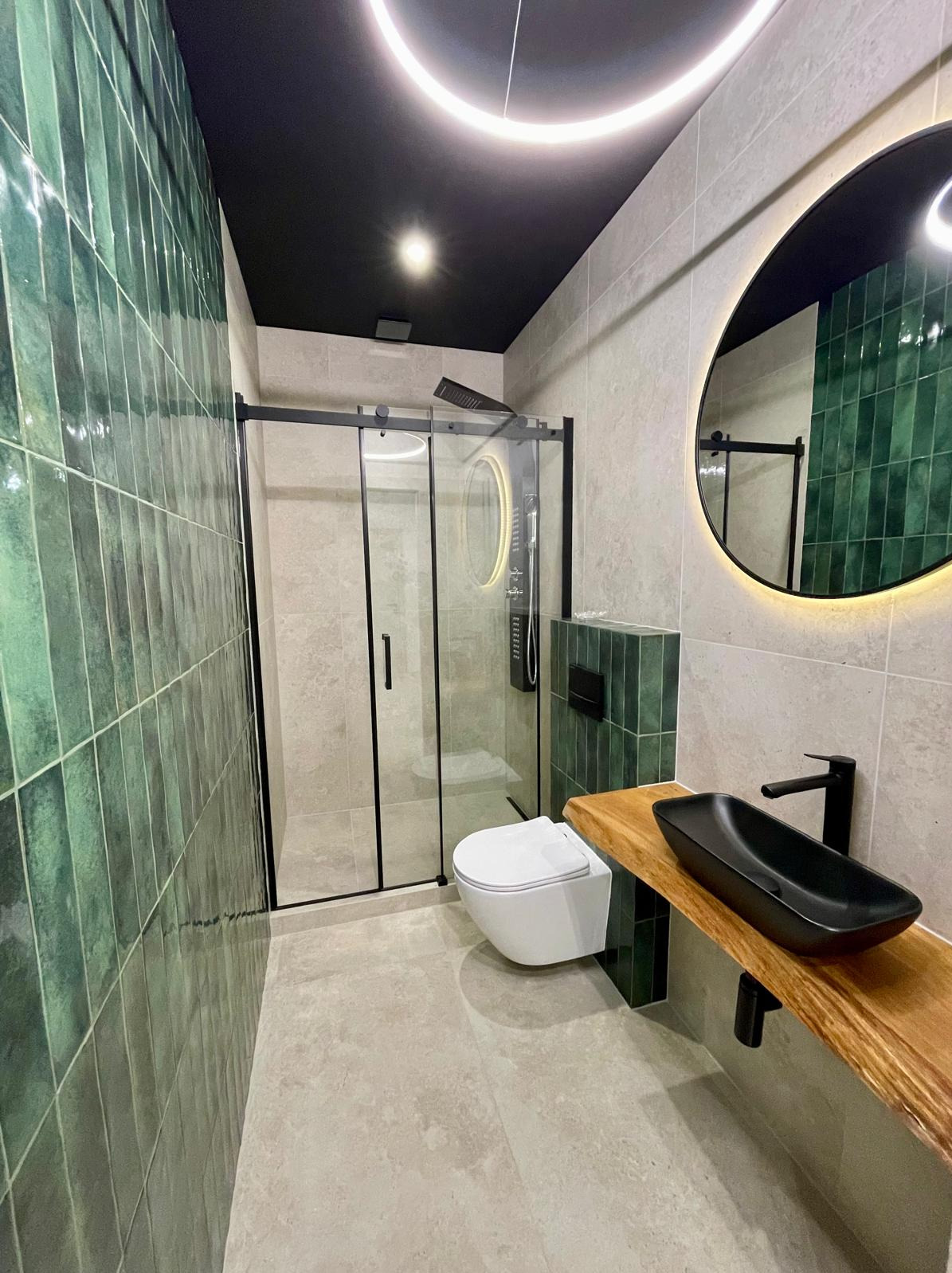 Nowoczesna łazienka z zielonymi płytkami, czarną armaturą i okrągłym lustrem z podświetleniem LED. Kabina prysznicowa z czarnymi ramami, umywalka nablatowa na drewnianym blacie.