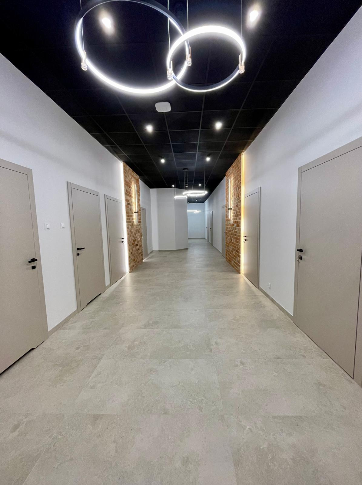Długi korytarz biurowy z szarymi drzwiami i ceglanymi wstawkami, oświetlony nowoczesnymi lampami wiszącymi w kształcie okręgów. Ciemny sufit i jasna podłoga.