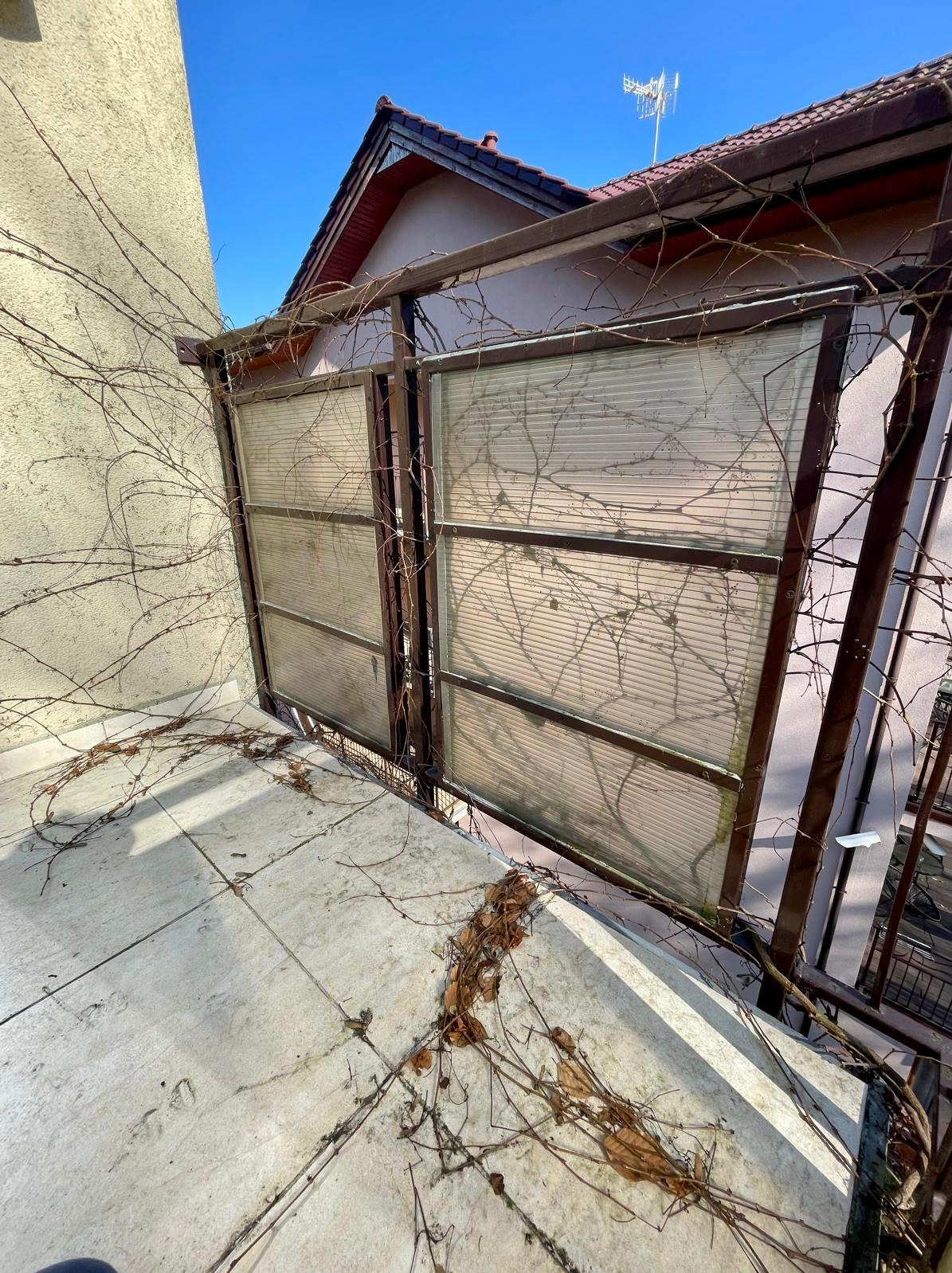 Zabrudzony balkon z zadaszeniem, pokryty suchymi liśćmi i gałązkami. Widoczne zacieki na płytkach i zabrudzone panele zadaszenia. Słoneczny dzień.