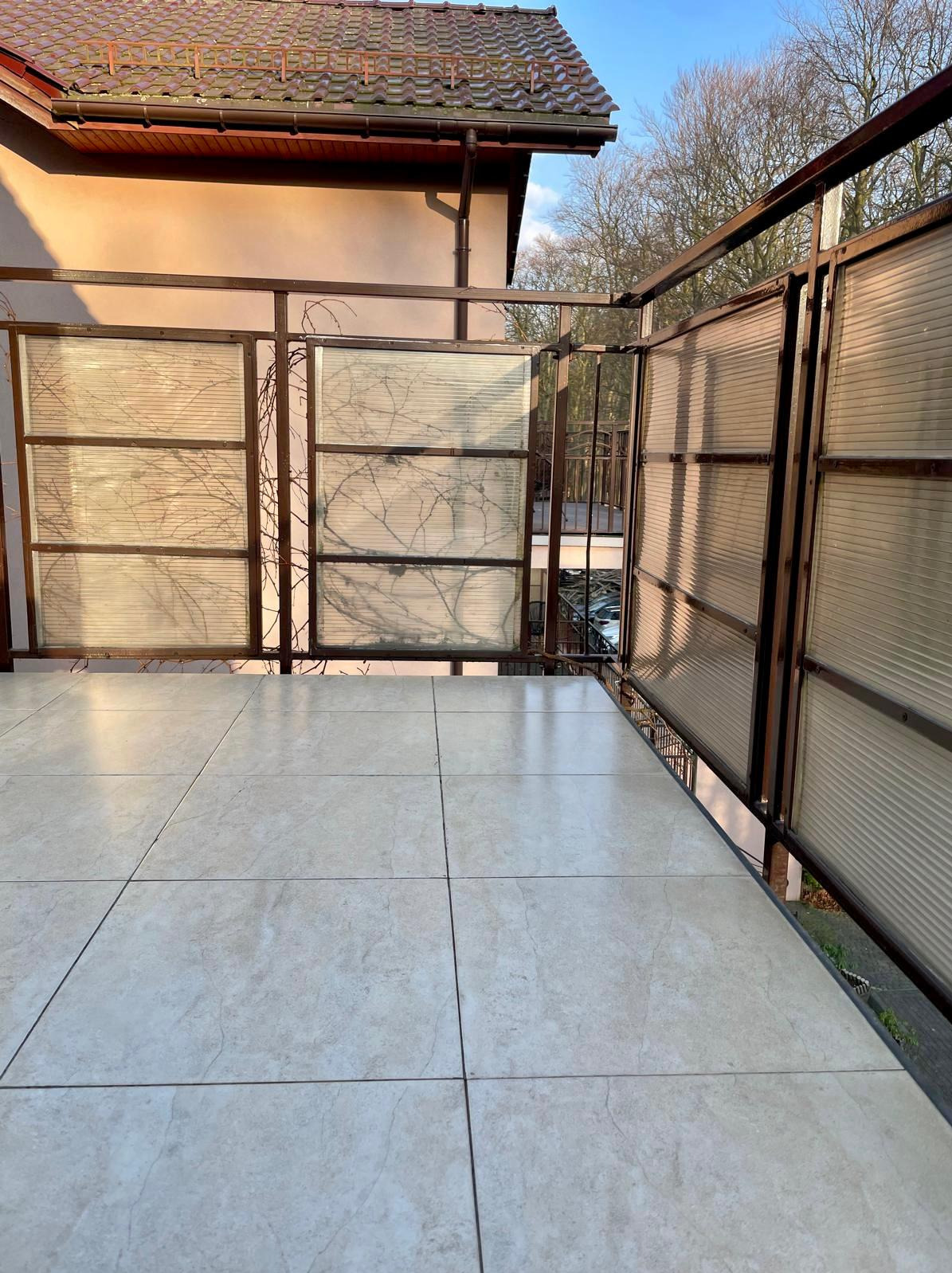 Jasne, ceramiczne płytki na balkonie z metalową balustradą i panelami z tworzywa. Widok na drzewa i budynek w tle. Słoneczny dzień.