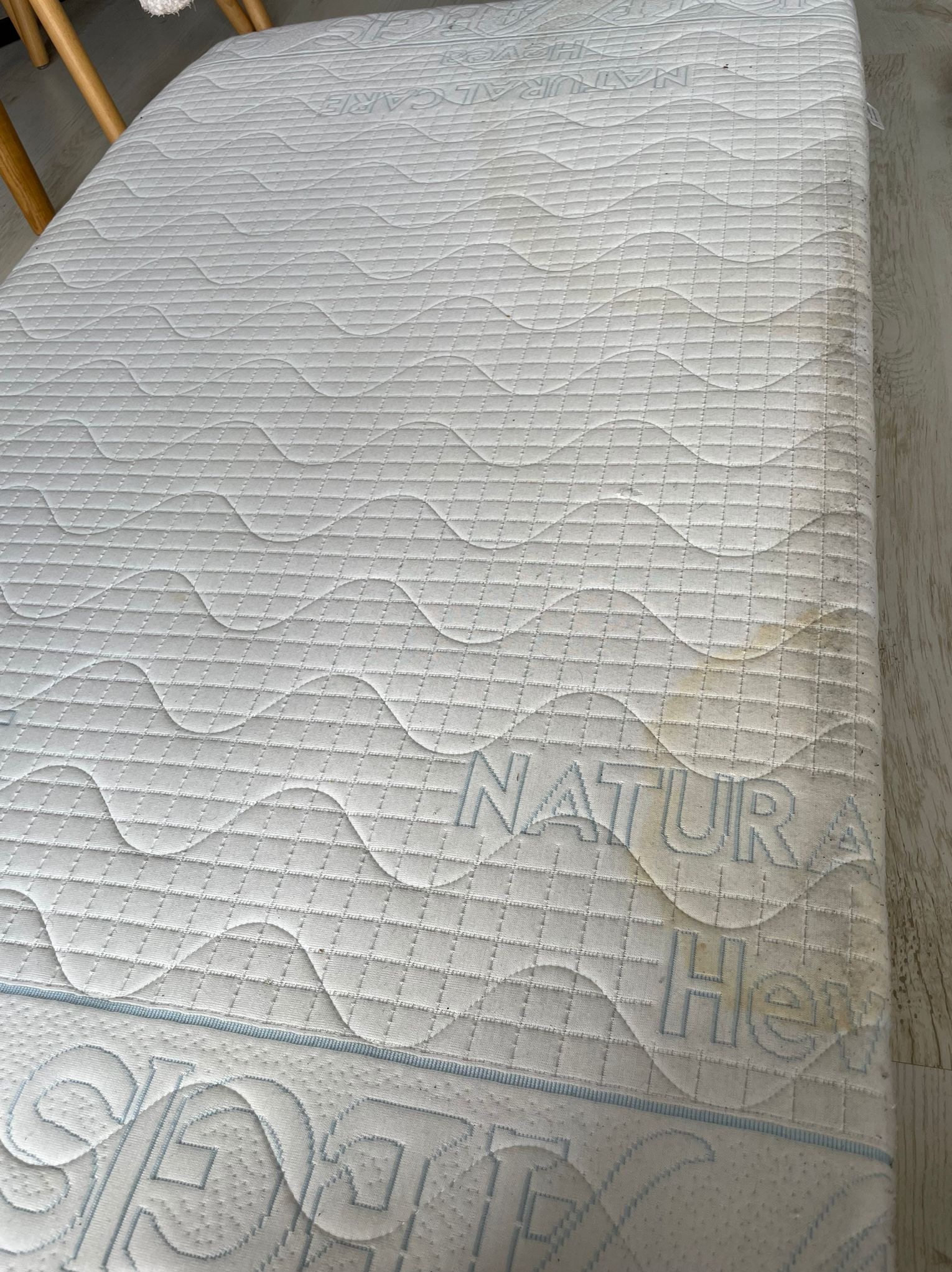 Materac z widocznymi plamami i zabrudzeniami na białym, pikowanym poszyciu. Widoczne napisy 'NATURA' i fragment 'SAFE'.